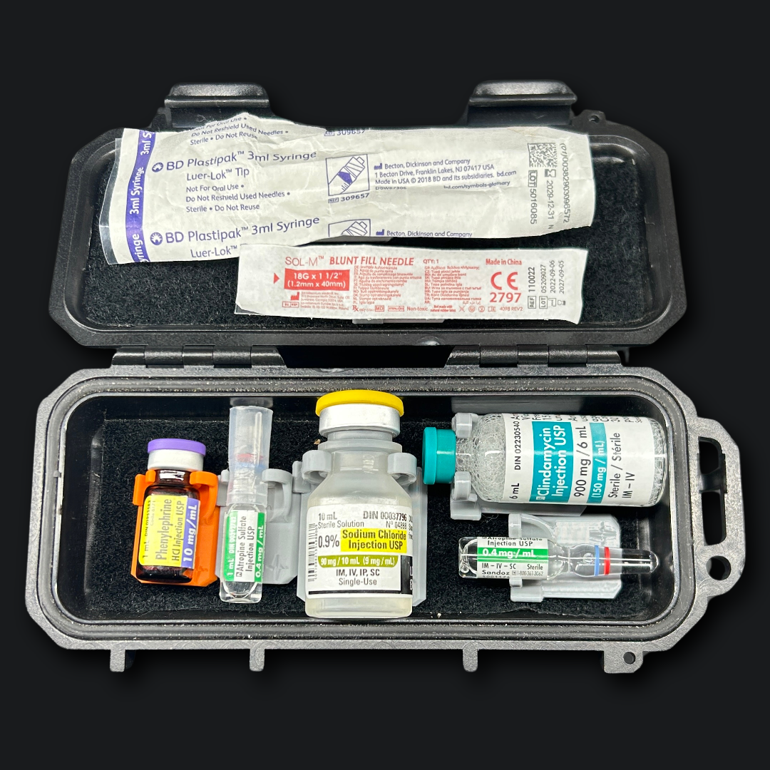 An open gray hard case containing medical supplies, including syringes, vials, a tourniquet, and packaged wound care items, photographed on a black background.