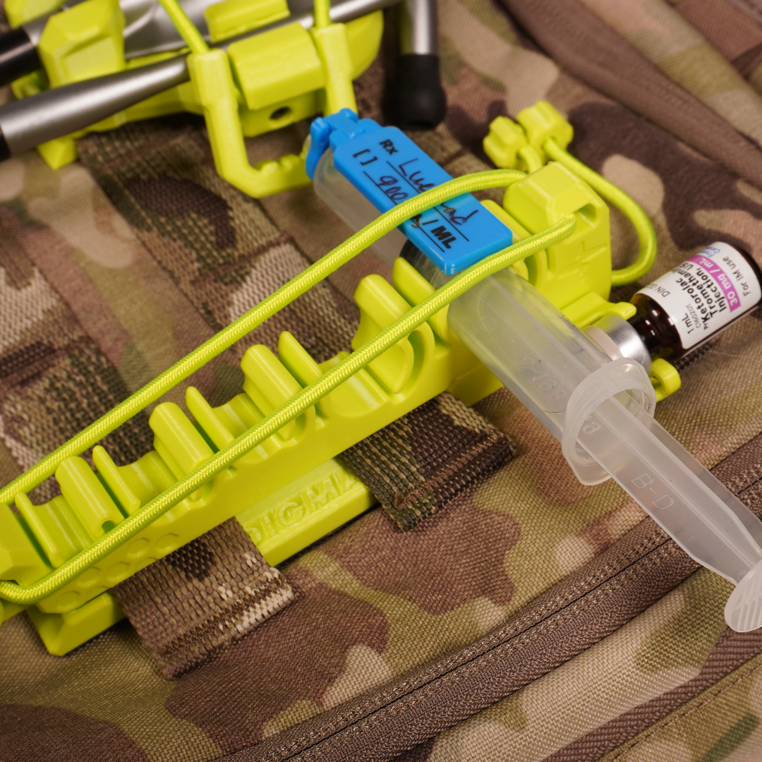 A close-up of a bright yellow-green plastic syringe holder mounted on a camouflage tactical backpack, holding a clear syringe with a small dark bottle.